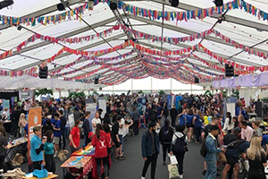 An image depicting the inside of a marquee with rows of tables parallel to one another and students visiting them and chatting to stall holders.